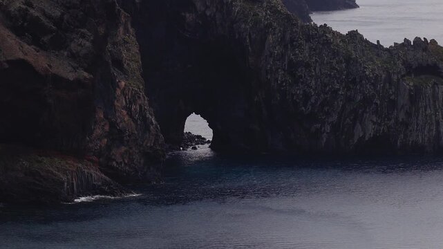 Aerial drone pass shows a sea arch in dark basalt cliffs on Madeira island, Portugal. Gentle waves lap the inlet as cool light shifts over deep blue water.