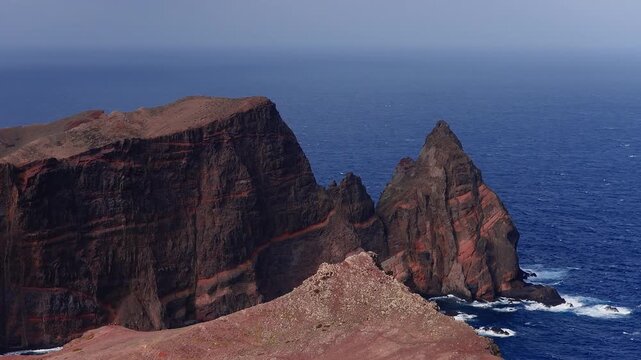 Aerial view of red banded volcanic cliffs at Ponta de Sao Lourenco, Madeira, Portugal, as waves crash below. Midday light, gentle pan and pullback show scale.