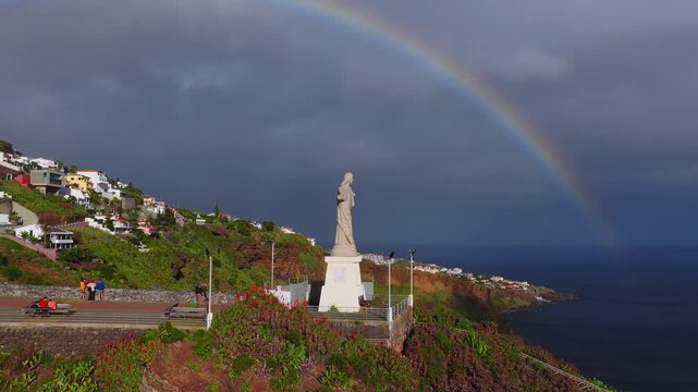 Aerial view shows Cristo Rei above the Atlantic near Funchal, Madeira. A double rainbow appears as people sit, stroll, and a runner passes on the promenade.