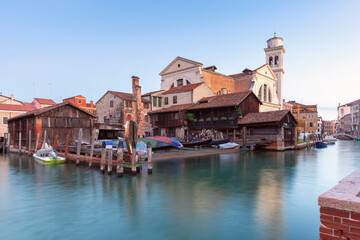 Traditional gondola workshop by canal with boats and church tower in Venice Italy at dusk © Kavalenkava