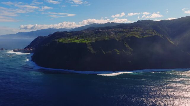 Aerial drone move over Madeira, Portugal reveals sea cliffs, terraced fields, ravines, low clouds, and Atlantic surf, with bright sunlight and soft haze in cinematic framing.
