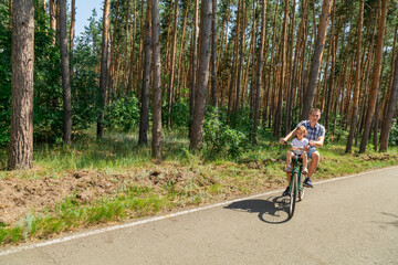 Naklejka premium father riding a bike in summer forest with his baby daughter sitting in child seat