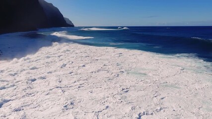 Aerial view of towering sea cliffs on Madeira, Portugal, as large Atlantic breakers roll in. Thick white foam races to shore, cliffs cast long shadows, colors are saturated.