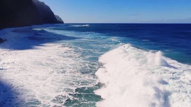 Aerial view over Madeira Island, Portugal, shows dark sea cliffs, powerful swells, and white breakers in midday light, with turquoise to navy waters and long shadows.