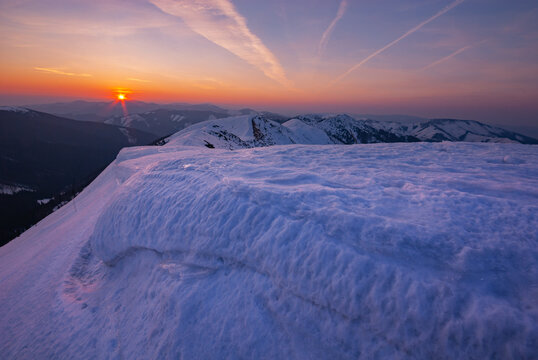 View of the sun setting behind snow-covered mountain peaks, casting an orange glow across the pristine white landscape, Rovna hola, Banskobystricky kraj, Slovakia.