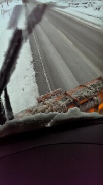 Cabin view of a motor grader plowing snow, with snow flying off the blade during winter road maintenance operations.