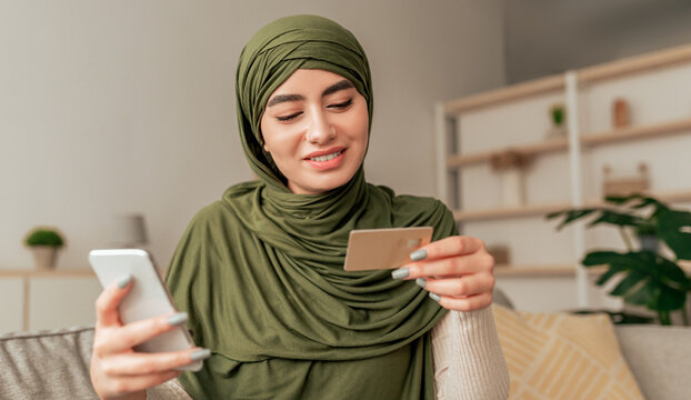 A young woman in a green hijab sits comfortably on a sofa at home, smiling as she checks her credit card while using her smartphone for online shopping. - Powered by Adobe