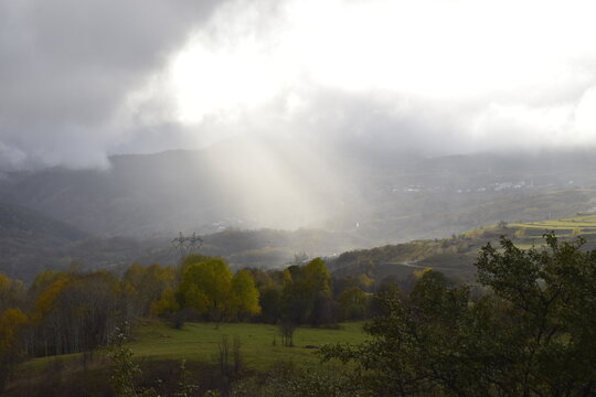 View of golden light piercing through heavy clouds, illuminating the serene landscape of rolling hills and autumn trees, Posof, Ardahan, Turkey.