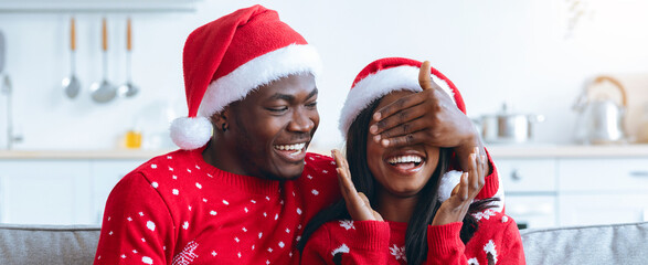 A joyful couple in matching Christmas sweaters and Santa hats laugh together on a couch. The woman playfully covers the man's eyes, creating a fun and loving atmosphere in their cozy home.