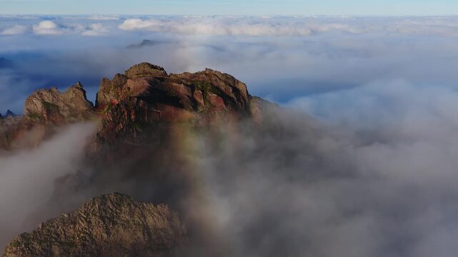 Aerial over Pico do Arieiro ridge in Madeira, Portugal. Rocky cliffs rise through clouds as mist swirls, a faint fogbow appears, and warm low light grazes textures.