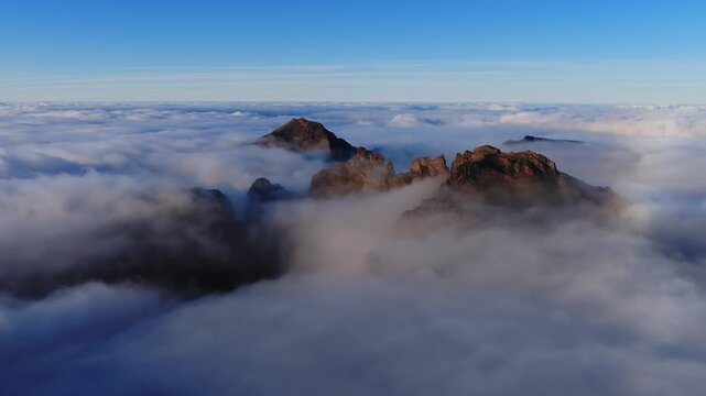 Rugged volcanic peaks in Madeira central highlands pierce clouds as early light warms rocky crags. Pico do Arieiro wide panoramic aerial view shows flowing mist and vertical relief.