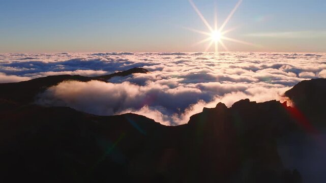 Aerial drone pan above cloud inversion at Pico do Arieiro, Madeira, Portugal. Mist pours through ridges, radar dome near summit, sunstar light and lens flare visible.