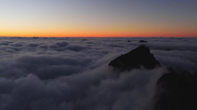 Aerial view shows jagged volcanic Pico do Arieiro above clouds at dusk in Madeira, Portugal. The camera glides past silhouetted summits and distant islets under pastel light.