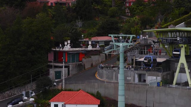 Aerial view of Funchal, Madeira shows cable car gondolas gliding above terracotta roofs, a yellow van turning, and visitors on terraces in soft overcast light.