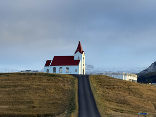 A serene Icelandic landscape featuring the red church on a gentle hillside, framed by vibrant grasslands and the striking white mountains of the Sn&aelig;fellsnes Peninsula.