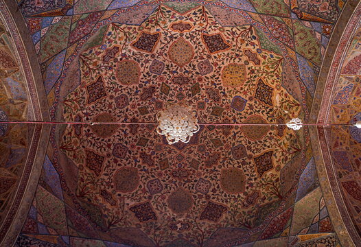 View of an intricately painted ceiling with floral and geometric patterns surrounding a chandelier, showcasing vibrant colors and textures, Lahore, Punjab, Pakistan.