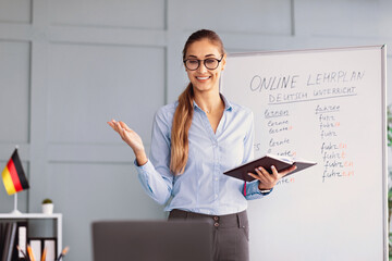 A female teacher stands in front of a whiteboard with German language materials while explaining a...