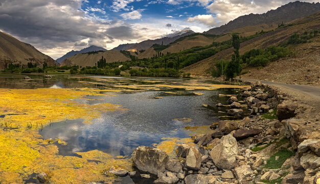 View of the serene lake reflects the cloudy sky and the rugged mountains, with yellow vegetation contrasting the water's blue, Gupis, Gilgit Baltistan, Pakistan.