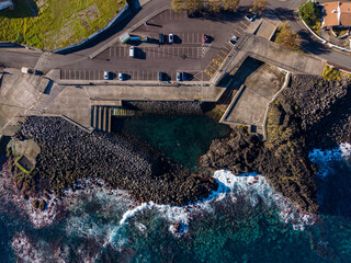 Aerial view of dark volcanic rocks embracing the turquoise waters near the concrete structures of Capelas, Azores, Portugal.