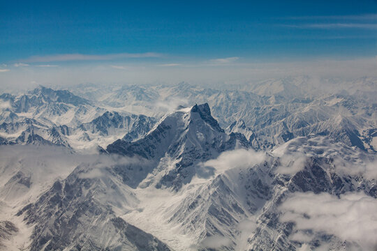 Aerial view of rugged, snow-capped mountains piercing through a sea of clouds under a serene blue sky, Skardu, Gilgit Baltistan, Pakistan.