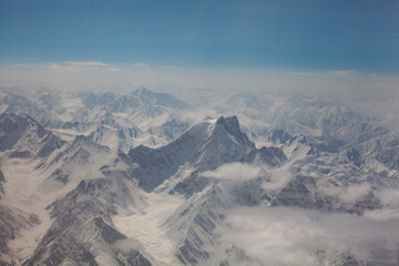 Aerial view of snow-capped mountains pierce the sky, their jagged peaks softened by ethereal clouds and a hazy blue atmosphere, Skardu, Gilgit Baltistan, Pakistan.