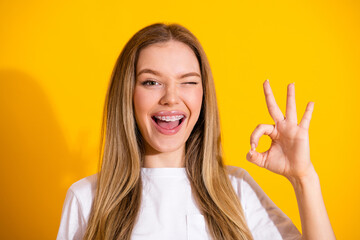 Young woman with braces winks and shows an ok sign against a bright yellow background wearing a...