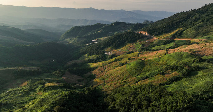 Aerial view of verdant hillsides cascading into shadowed valleys, a tapestry of green and brown under a soft sky, Tambon Tap Tao, Chang Wat Chiang Rai, Thailand.