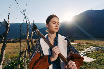 Fototapeta premium woman outdoors mountains sunrise rural stick jacket nature A young woman in a warm jacket holds a wooden stick in a sunlit field, eyes closed and serene against distant mountain peaks.