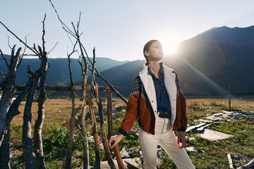 Fototapeta premium Woman in patterned jacket standing in a field with mountains and warm sunlight, posing near rustic wooden sticks and rocks, fashion portrait and lifestyle in natural outdoor landscape.