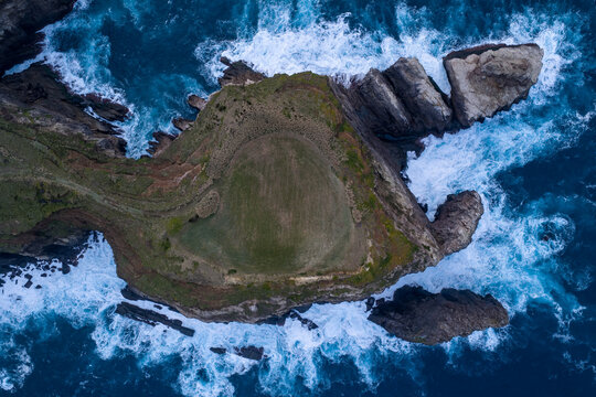 Aerial view of a rugged, verdant island, a stark contrast to the tempestuous azure sea crashing against its rocky shores, Ribeirinha, Azores, Portugal.