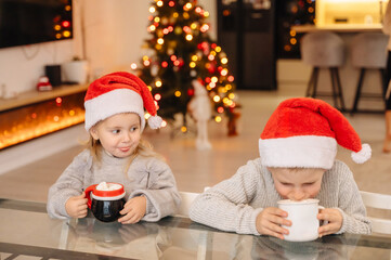 Two children in Santa hats happily drinking a hot beverage from mugs by the Christmas tree, holiday tradition