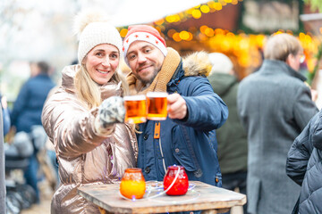 Happy couple drinking warm punch and mulled wine with a Christmas market full of lights shining...