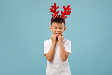 A joyful young boy stands against a blue background, wearing vibrant reindeer antlers. He closes...