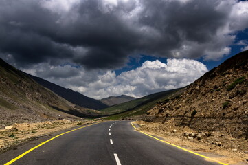 View of a winding asphalt road cutting through a valley, contrasting with the rugged, rocky terrain under a dramatic, cloudy sky, Naran, Khyber Pakhtunkhwa, Pakistan.