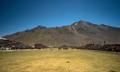 Shandur, Pakistan - 31 July 2017: View of a vast, sun-drenched polo field teeming with spectators against the backdrop of rugged mountains and a clear, azure sky.