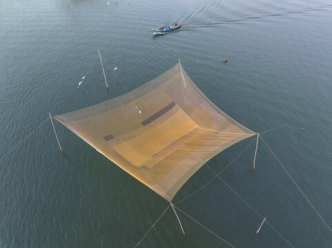 Aerial view of a vast, sun-drenched fishing net shimmers like gold against the deep turquoise waters, while a distant boat glides by, Hội An, Quảng Nam, Vietnam.