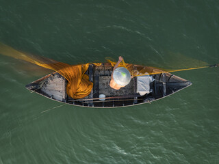 Aerial view of a lone fisherman casting a vibrant yellow net from his weathered boat on the rippling green waters, Hội An, Quảng Nam, Vietnam.