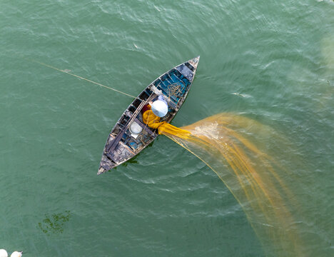 Aerial view of a lone fisherman casts his vibrant yellow net from a weathered boat, creating a striking contrast against the serene turquoise waters, Hội An, Quảng Nam, Vietnam.