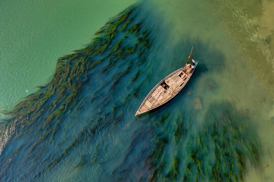 Aerial view of a rustic wooden boat cutting through the tranquil, algae-streaked waters, creating a vibrant contrast against the sandy bank, Sirajganj, Rajshahi Division, Bangladesh.