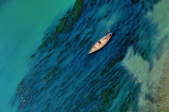 Aerial view of a lone wooden boat sailing through the river, its weathered texture contrasting with the vibrant turquoise water, Sirajganj, Rajshahi Division, Bangladesh.