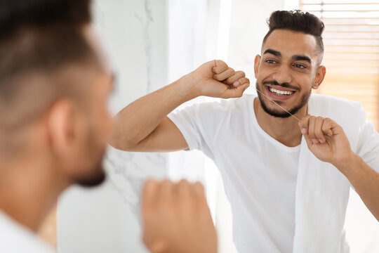 A young man brushes his teeth and flosses in a bright bathroom. He appears cheerful while looking in the mirror, focusing on his dental care.