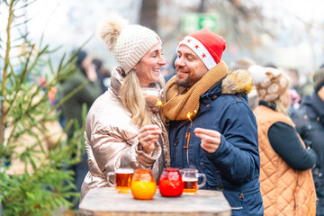 A cheerful couple holding punch glasses and sparkling sparklers in front of a festive Christmas market backdrop in Bavaria Germany on a cozy winter night