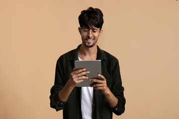 A young man stands in a neutral colored environment, smiling as he interacts with a tablet. He wears glasses and appears engaged and happy during the day.