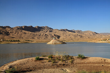 View of tranquil blue waters reflecting the clear sky, with a small island topped by a white structure, all framed by rugged, sun-drenched mountains, Quetta, Balochistan, Pakistan.