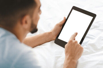 A young man sits comfortably on a bed, using a tablet. He is focused on the screen, tapping with his finger. Soft lighting creates a calm atmosphere in the bedroom.