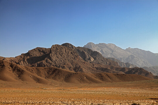 View of rugged, sun-baked mountains rise dramatically against a clear blue sky, casting long shadows across the arid landscape, Quetta, Balochistan, Pakistan.