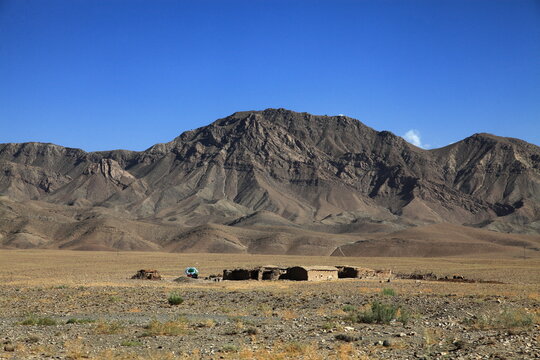 View of arid plains stretching to meet the rugged, textured mountains under a vast blue sky, with a small building nestled in the foreground, Quetta, Balochistan, Pakistan.