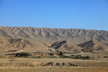 View of arid mountains rise dramatically behind a sparse village with muted buildings and scarce greenery under a clear blue sky, Quetta, Balochistan, Pakistan.