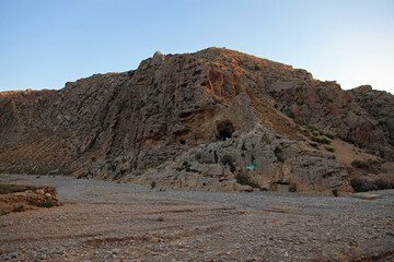 View of a rugged, sun-baked mountain looms, its rocky face scarred with caves and crevices, overlooking a parched, stony ground, Quetta, Balochistan, Pakistan.