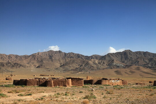 View of sun-baked mud structures stand stark against a backdrop of rugged, arid mountains under the vast, cloud-dotted sky, Quetta, Balochistan, Pakistan.